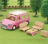 Pink toy car with rooftop tent, outdoor furniture, and picnic setup on a grassy area with trees and flowers.